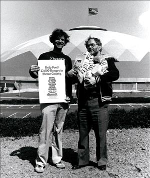 Two people standing in front of a dome shaped stadium. One is holding boxes and cans of food. The other is holding a sign that says help feed twenty two thousand in pierce county. 